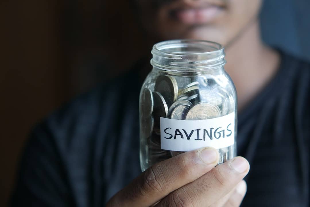 Ive people successfully climbing a mountain made of coins, each holding different budget-friendly items like a piggy bank, calculator, shopping bag with sale tag, a grocery list, and a budget chart